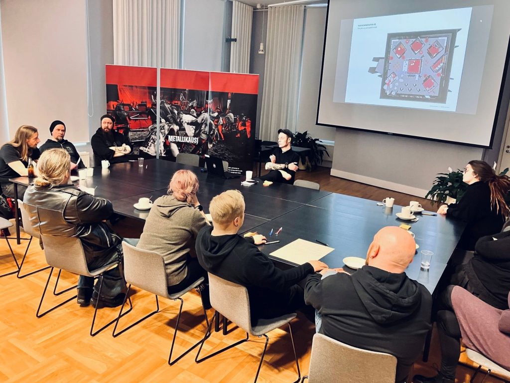 Noin kymmenen henkilöä istumassa pöytäryhmässä. Taustalla näkyy heijastettuna näyttelysuunnitelma ja nurkassa kolme Metallikausi roll-upia.
