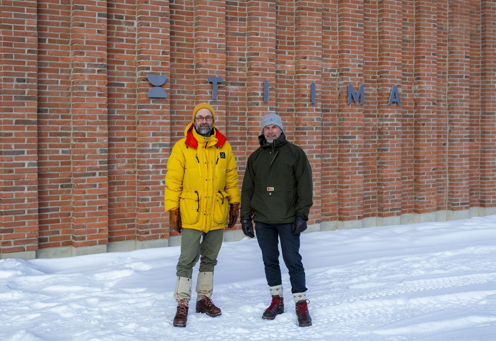 Kaksi henkilöä seisomassa talvivaatteissa tiiliseinän edessä.