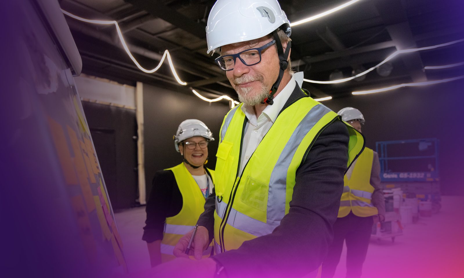 Kolme työmaavarusteisiin pukeutunutta henkilöä Tiiman työmaalla. Yksi henkilöistä nautaa iloisesti ja toinen tekee jonkinlaista tehtävää fläppitaululla.