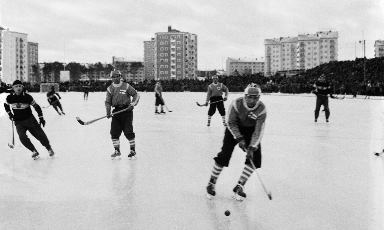Mustavalkokuva, jääpallon pelaajia jäällä, taustalla kerrostaloja.