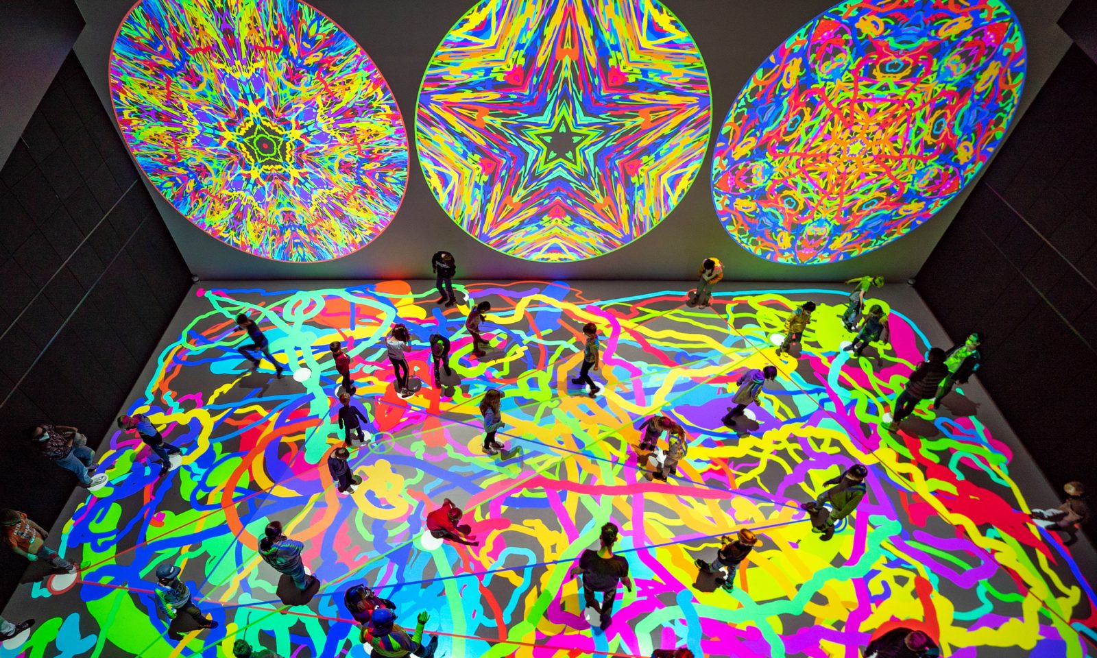 People in an immersive space, with colourful shapes projected onto the walls and ceiling.