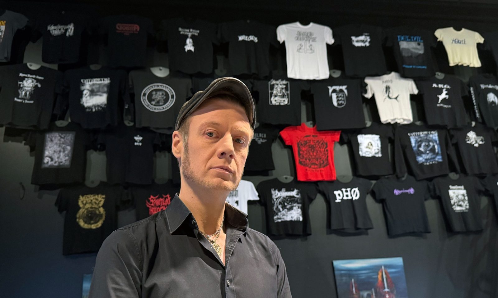 Henkilö seisoo Metallikausi-näyttelyn bändipaitaseinän edessä.